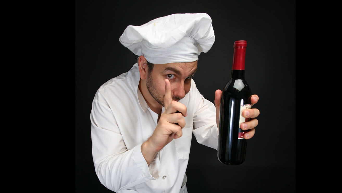 Férfi szakács feltartott ujjal, másik kezében egy zárt vörösboros üveg fekete háttér előtt