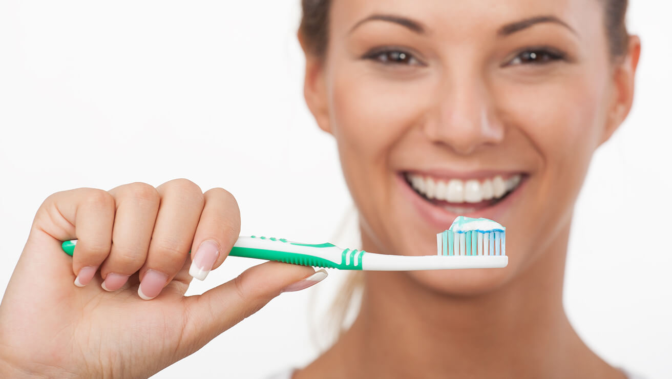 Mosolygó nő tartja a zöld-fehér fogkefét rajta fehér-kék-zöld fogkrémmel fehér háttér előtt