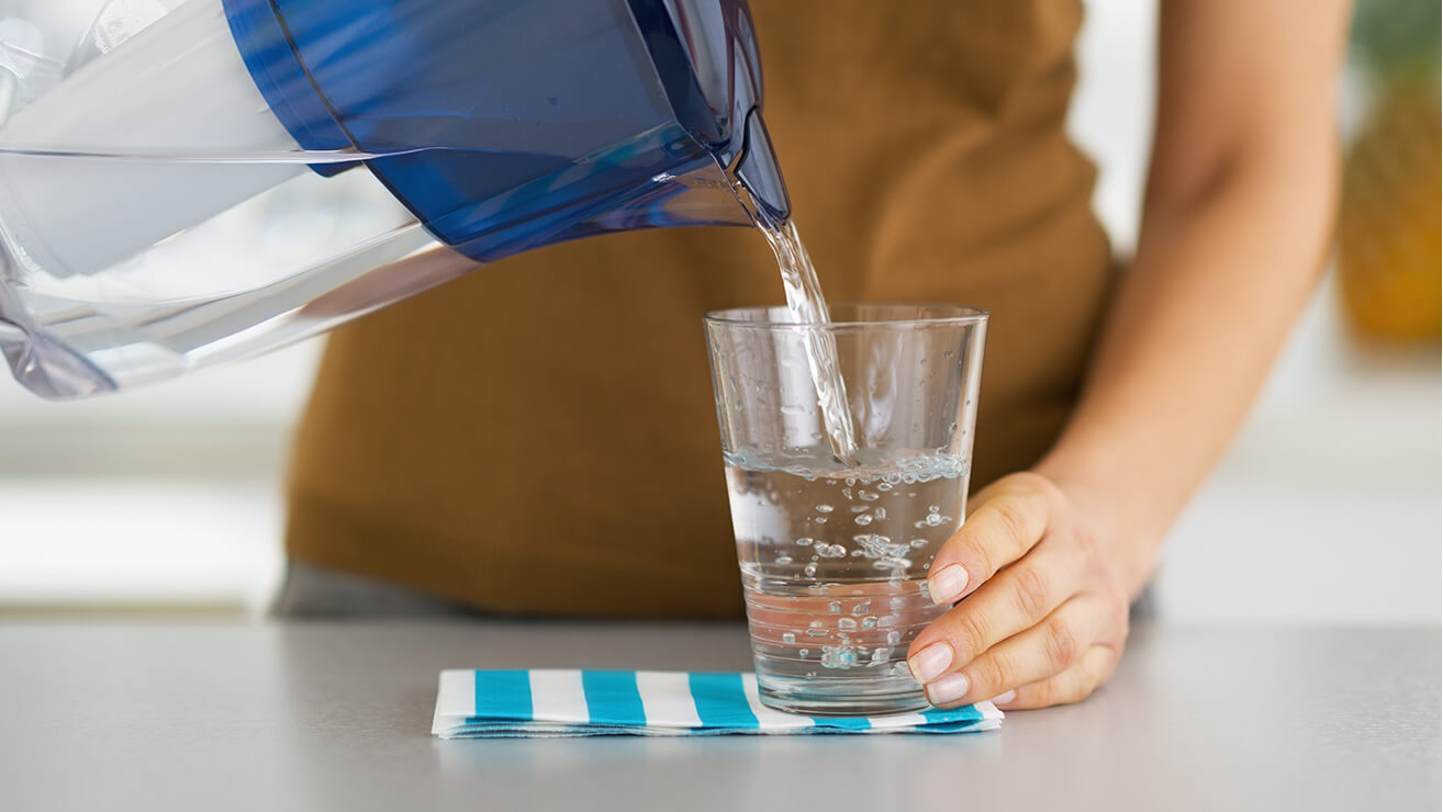 Nő vizet önt egy vízszűrős kancsóból egy szalvétán álló pohárba a munkapulton
